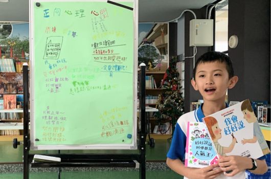 一個小男孩穿著藍白色T恤，在室內舉著一份文件，身旁是一塊綠色的立式白板，上面用紅筆和黑筆寫滿了中文字和數學算式，背景是家中書架和裝飾物品。