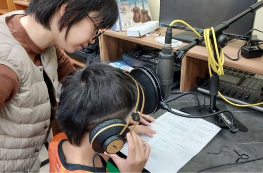 兩名學生在錄音室進行語音錄製活動。戴著耳機的學生坐在麥克風前閱讀稿件，身後的學生協助監控。桌上擺放錄音設備、麥克風、耳機和文件。