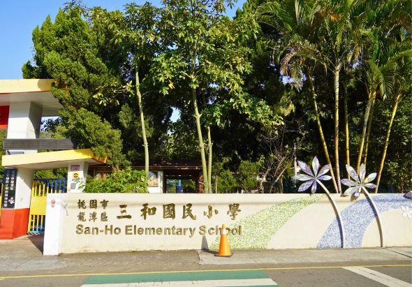 三和國小校門口，白色牆面上有中英文校名標示「三和國小」和「San-Ho Elementary School」，校舍為現代建築風格，門口兩側種植高大綠樹，校園綠意盎然，停車格標線清晰可見。