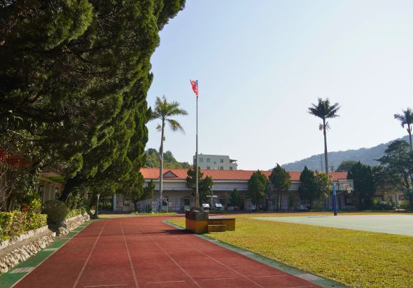 校園廣場景觀，紅色跑道路徑沿左側延伸，中央草坪開闊，遠處可見校舍建築、飄揚的紅旗、棕櫚樹和山景，整體環境整潔寧靜。