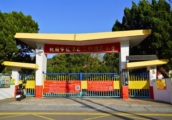 幼兒園大門，黃色屋頂配紅藍色牆面，門口掛著紅色標語布幔，周圍綠樹環繞，門前停放一輛紅色機車，晴朗天氣下的校園入口景觀。