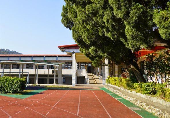 學校運動場景：紅綠相間的塑膠跑道與網球場，旁邊是現代化的校舍建築，屋頂為紅色鐵皮，白色牆面，周圍種植大型榕樹和綠色灌木，清晨陽光灑落，場景寧靜宜人。