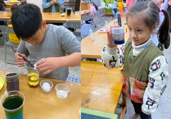 兩名小學生在教室進行科學實驗活動，桌上放有裝有深色液體的杯子、小白碗和其他實驗用具。左邊男生正用湯匙操作實驗，右邊女生觀察著實驗過程。背景可見其他學生和老師。