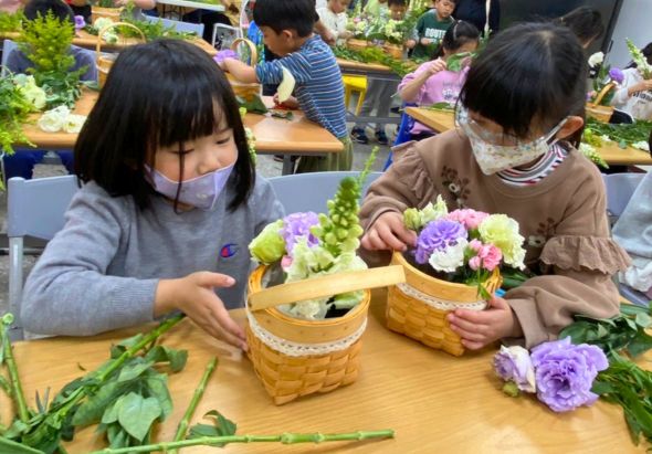 一個教室內，兩名戴著口罩的小女孩坐在桌子前，正專注地將紫色和粉色的花朵插入竹編花籃中。桌上散放著綠色莖葉和花朵，背景可見其他學生也在進行相同的插花活動。