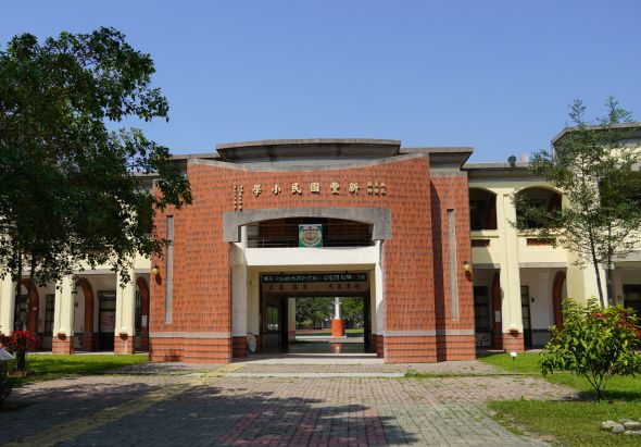 紅磚建築的博物館正門，門口上方有中文標誌，前方有磚砌地面，兩側種有綠樹和植物，建築風格簡潔現代，晴朗藍天背景。