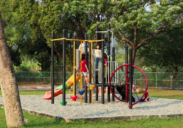 公園遊樂場景，中央為多功能組合遊具，包含深灰色主塔、黃色溜滑梯、紅色環形攀爬架及其他攀爬設施。周圍鋪設碎石地面，被綠樹環繞。數名兒童穿著紅、黃、白色衣物在遊具上玩耍。背景有金屬圍籬和草坪。