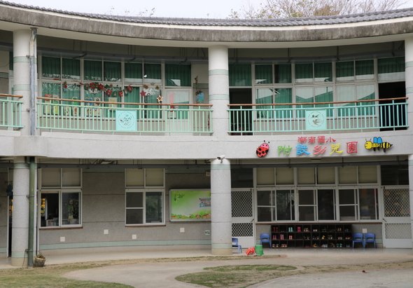 一棟幼稚園或托兒所的建築物，由兩層樓組成。上層有綠色欄杆的露台和多扇窗戶，下層是教室和活動區域。牆上貼有彩色兒童教育圖案和文字標誌。地面放置玩具和教具。