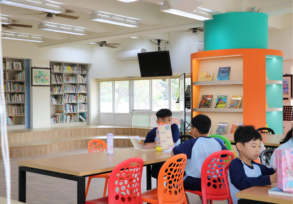 現代化學校圖書館內，四名學生坐在木製長桌前學習，桌上擺放紅色和橙色塑膠椅。背景有木質書櫃、彩色儲物架、落地窗和天花板照明設備，營造明亮開放的學習環境。