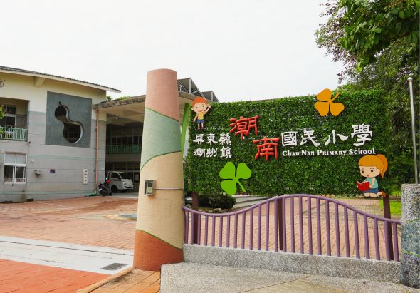 照片為潮南國小校門入口，以綠色植栽牆面為背景，展示校名招牌及彩色花卉裝飾。前方有紅色欄杆，校舍建築物可見於左側。