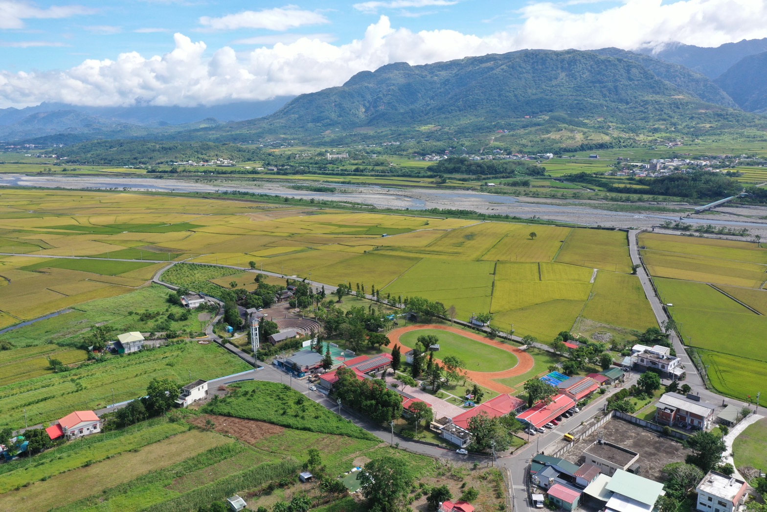 俯視遼闊的農業谷地，綠色稻田與河流交錯，中央有紅色跑道運動場與校舍建築。背景是翠綠山巒，遠處村落散佈，天空雲朵飄浮。