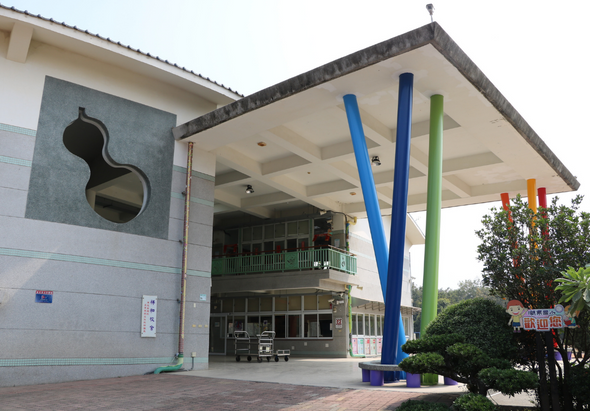 現代建築外觀，白色牆面搭配灰色屋頂，左側牆上有黑色藝術裝置，右側有藍、綠、黃、橙色豎立柱子，寬敞的開放式入口，前方有綠化植栽，右側可見紅色招牌。