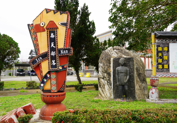 戶外公園景觀，左側為紅黃色傳統風格的大型燈籠雕塑，刻有中文字樣；右側為石頭雕刻的人物雕像。周圍種植綠樹和灌木，背景可見建築物和停車場，展現傳統文化主題的公共裝置藝術。
