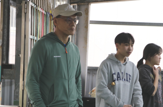一名戴著棒球帽的男性導師穿著綠色夾克站在左側，三名年輕學生站在右側，其中兩人穿著灰色衛衣。他們在室內空間中，背景可見窗戶和籠子。場景呈現教學或指導活動的氛圍。
