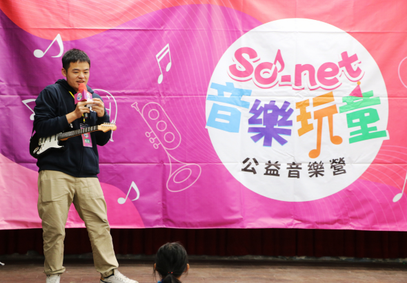 一位年輕男性在舞台上手持麥克風和吉他進行表演，身穿黑色外套和米色褲子。身後為粉紅色布景，上有音樂符號圖案。右側白色圓形標誌寫著「SoNet音樂玩樂」和「公益音樂營」的中文文字。舞台下方有觀眾坐著。