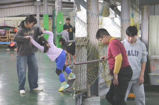 室內運動館內，一位成人協助穿著粉紅上衣的幼童進行吊環運動，幼童身體倒掛。其他幾位穿著深色衣服的大人和穿著校服的學生在一旁觀看或等候。背景有綠色運動器材和安全網。