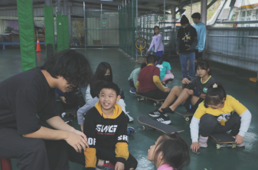 室內體育館中，一位穿黑衣的教練正向坐在地上的兒童們教授動作。孩子們穿著黃黑相間的運動服，認真跟隨指導。背景有其他參與者和工作人員，顯示這是一場兒童運動培訓或課程活動。