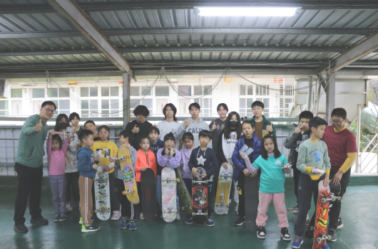 一群大人和小孩在室內運動場集合，許多孩子拿著滑板，穿著各色運動服裝，在成人指導下參與滑板培訓或教學活動，背景是鐵皮屋頂的場地。