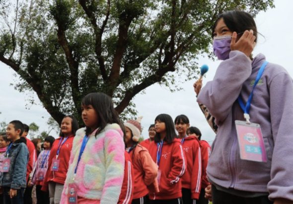 戶外活動中，一位戴著紫色口罩、穿著淺紫色衣服的女性講者手持麥克風，面向穿著紅色運動服的學生群體致詞。大樹下聚集了多名配戴識別證的參與者，氣氛熱烈。