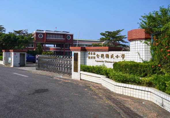 學校校門口景象，白色磁磚圍牆上有校名標誌，內側為紅磚建築校舍，旁邊有紅色水塔，鐵製推拉門關閉，周圍種植綠樹，晴朗天氣。