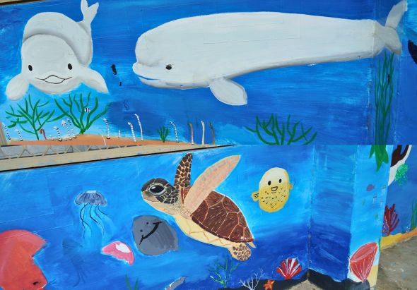 海洋生物主題壁畫，描繪白色鯨魚、海龜、海豚、海蜇、珊瑚礁及各式彩色魚類在藍色海洋中悠游的場景，色彩鮮豔，適合兒童教育展示。