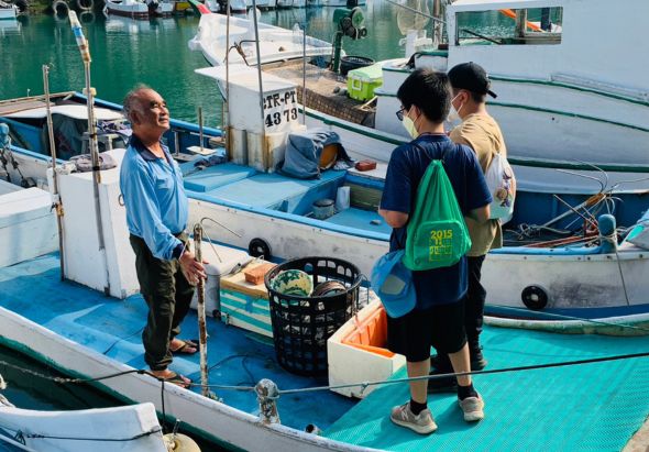 漁港碼頭上，一名漁民正向三位遊客介紹漁船和漁業工作。背景停泊著多艘藍色漁船，前景展示漁具和捕獲物。遊客穿著休閒服裝，認真聆聽解說，體驗傳統漁業文化。