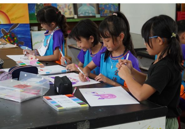 四名穿著紫色和藍色制服的小學女生在教室內圍坐在黑色桌子前進行美術創作。她們專注地用彩色筆在白紙上繪畫，桌上放著色筆、調色盤等美術用具。背景牆面掛有學生作品展示。
