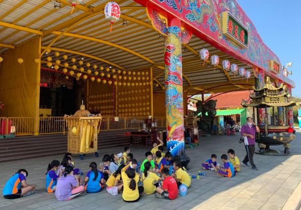 寺廟前廣場上，身穿藍黃色衣服的兒童們聚集坐在地上，參與宗教活動。背景為金黃色支架和紅色廟宇建築，裝飾著紅燈籠與彩繪柱子，展現濃厚的亞洲傳統宗教氛圍。