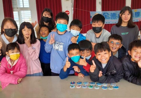 一群小學生在教室內合影，多數學生戴著彩色口罩，圍繞著桌前展示的手工藝品。背景有紅色窗簾和牆上的海報。學生們做出開心的手勢，呈現溫馨的集體活動氛圍。