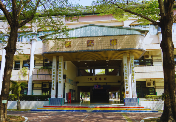 校園建築正面，具現代風格的入口大廳，白色與黃色外牆，大型玻璃窗戶，兩側高大樹木遮蔭，入口處紅藍色地面標線，整體建築呈對稱設計。