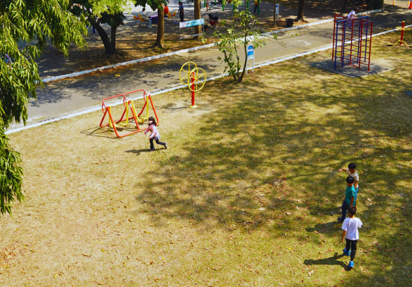 公園內，兒童在沙地上玩耍。左側有紅黃相間的攀爬架和轉盤等遊樂設施，右側有兩名兒童站立。背景有樹木、步道和其他遊樂設備，陽光灑落形成樹影。