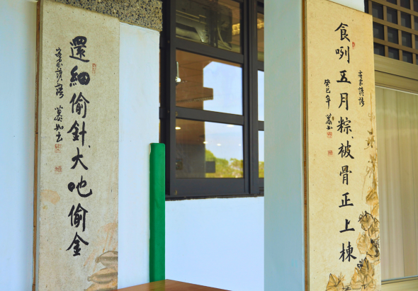 建築物入口處展示兩幅古文書法對聯，左幅寫有「農島倚針大也俞合」，右幅寫有「食吳五月粽被骨正上棋」，均為黃色底布上的黑色毛筆字，中央為玻璃門窗，體現傳統文化氛圍。