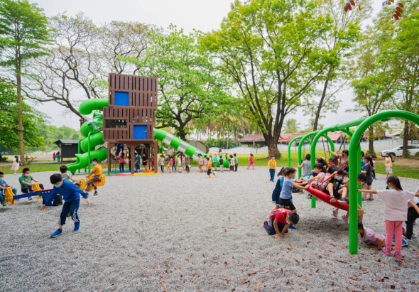 公園兒童遊樂場景，多名小朋友在沙地上玩耍。中央有棕色木製大型遊樂設施，周圍綠色攀爬架和滑梯。許多穿著黃色和藍色衣服的孩童四處活動，大樹提供遮蔭，營造熱鬧歡樂的戶外遊戲環境。