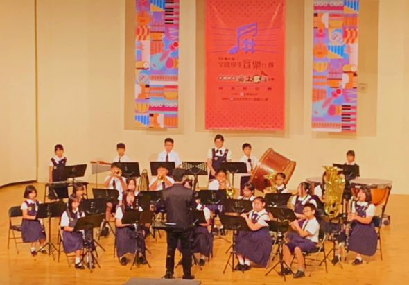 學生樂團在舞台上進行音樂演奏。約二十位穿著紫色制服的學生演奏各種樂器，由指揮者帶領。舞台背景掛著三幅彩色海報，牆面為米色。