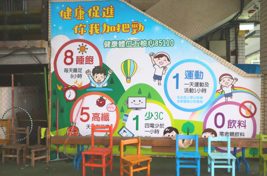 健康促進海報展示兒童健康生活習慣，包含8小時睡眠、5份高纖、1小時運動、少3C和0飲料等五大目標。彩色圓形圖示搭配卡通人物和相關圖案，下方擺放多色兒童椅子。