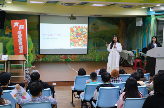 室內活動現場，講者在綠色植物背景前進行演講。投影幕展示彩色花卉圖案，聽眾坐在藍色椅子上認真聆聽。左側豎掛紅色中文橫幅。舞台右側站著另一位主持人。