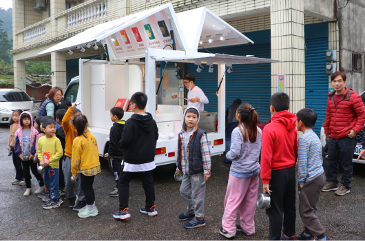一輛流動服務車停在建築物前，多名兒童排隊等待。車上有工作人員在服務窗口為民眾解說或提供服務。兒童穿著各色衣服，有的穿黃色、紅色或其他顏色的外套，聚集在車周圍參與活動。