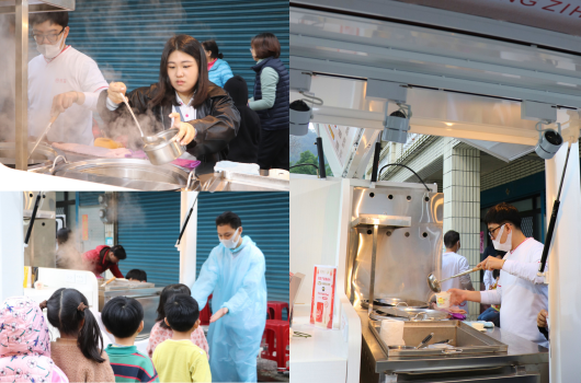 社區活動現場，多位志工和參與者在不同的攤位進行食物準備和烹飪示範。左上方女性在料理台前操作，左下方有小孩圍觀，右側有志工在廚房設備前忙碌。整個場景呈現温暖的社區互動和食物教育活動。