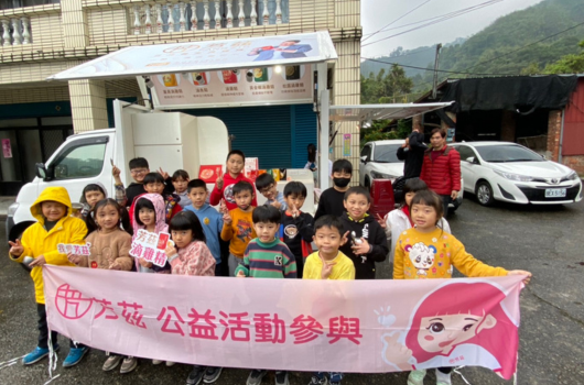 一群兒童與成人在停車場前合影，共同舉著寫有「女力展敵公益活動參與」的粉紅色布幔。參與者穿著紅黃色背心，背景為山景和建築物。