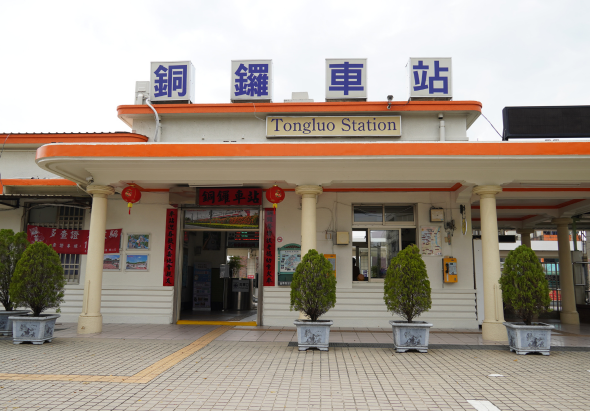 銅鑼車站外觀，建築以白色和橘色為主，正面掛著中英文站名招牌，門前擺放四盆綠色盆栽，左側懸掛紅色燈籠，地面鋪設淺色磁磚，整體設計典雅簡潔。