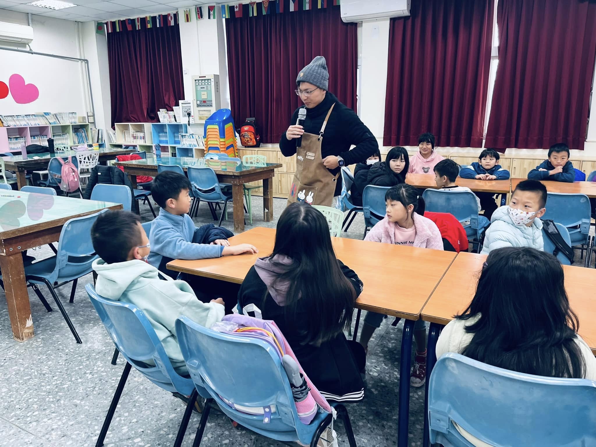 學校教室內，一位戴著灰色毛帽的女性講師手持麥克風，向圍坐成半圓形的約十五名小學生進行互動教學。教室背景有深紅色窗簾、國旗裝飾和書架。講師穿著棕色圍裙，學生認真聆聽。