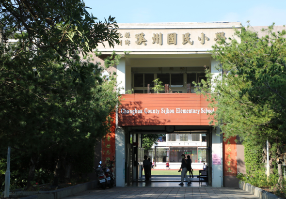 昌邑縣西洲小學校園入口，紅色招牌標示校名，白色建築主體，綠色植物環繞校門兩側，幾位人物在校門前活動，展現學校的校園環境與日常景象。