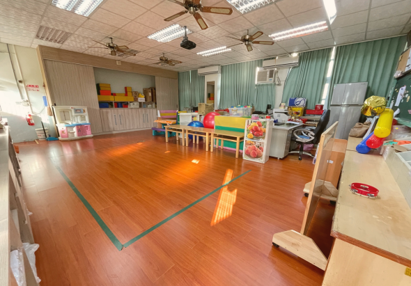 寬敞明亮的幼兒園教室，木質地板中央有綠色邊框標示區域。左側有木製收納櫃和黃色儲物盒，右側放置彩色小桌椅、教具和玩具。天花板裝有風扇和日光燈，綠色窗簾採光良好。空間整潔有序，適合幼兒學習活動。