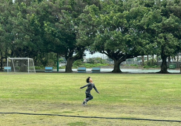 一位穿著黑色運動服的人在草地上奔跑訓練，身後是足球球門。公園內有大樹、河流和綠色草坪，環境開闊，適合戶外運動。