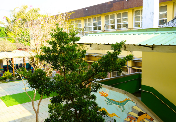 幼兒園校園場景，黃色建築物前方有綠色遮陽棚，院子裡種植綠色植栽，地面鋪設彩色壁畫，背景可見多層樓校舍和藍天白雲，環境明亮溫暖。