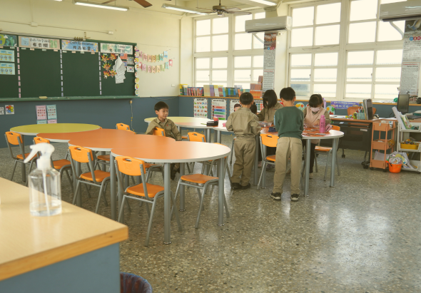 小學教室內，數名學童穿著校服在橙色桌椅旁學習。教室配備黑板、書架、教材展示牆和大型窗戶。地板明亮整潔，多張學習桌分散放置，呈現活躍的教學環境。