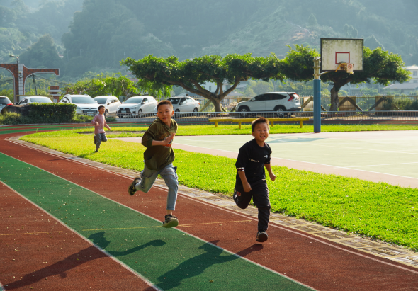三名兒童在室外運動場跑步，紅綠色跑道環繞綠色草地。背景可見籃球架、停放的車輛和大樹。陽光充足，景色優美，展現孩童運動與戶外活動的歡樂情景。