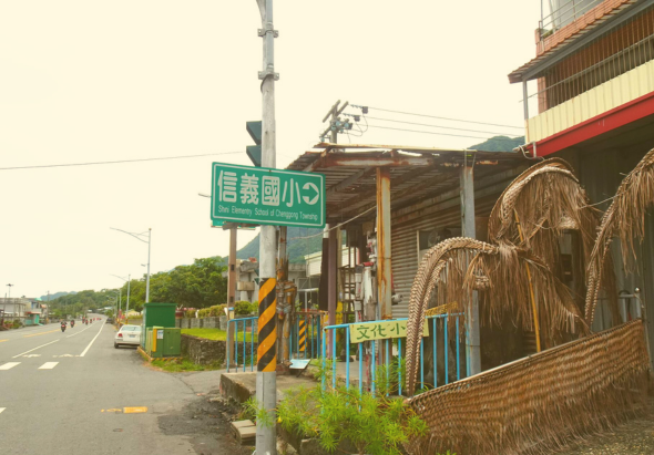 鄉村路邊景象，綠色路牌標示「信義國小」，右側為磚造建築，門前有彩色柵欄和垂柳裝飾，路上停有汽車，電線杆和交通標誌林立，整體呈現寧靜的小鎮風貌。