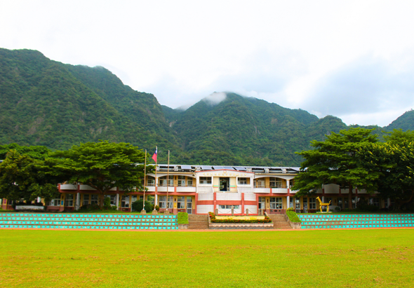 一所學校建築坐落於綠色草坪上，建物為橙白相間的三層樓房。背景為蒼翠的山脈和雲霧，前方為藍色的看台席。校園環境優美，群山環繞，樹木茂密。