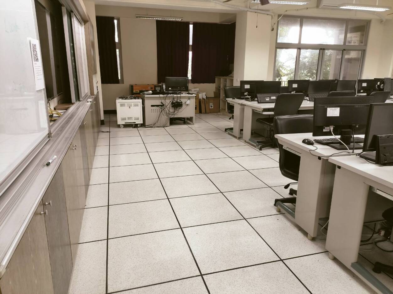 一間大學電腦教室，配置多台桌上型電腦和顯示器，白色地板鋪設黑色邊框瓷磚。左側有白板和儲物櫃，窗戶裝有深色窗簾。中央散置實驗設備和機器，右側排列多個工作站。天花板有照明設備，整體呈現空曠的教學環境。