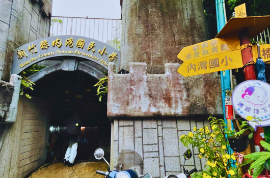 老舊建築入口處，拱形門洞上方掛有黃色中文招牌，門邊貼著黃色指示牌和彩色裝飾，門口放置掃帚等物品，牆面斑駁陳舊，爬滿綠色植物，呈現懷舊的街道風景。